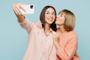Elder fun parent mom with young adult daughter two women together wear casual clothes doing selfie shot on mobile cell phone hug cuddle kiss isolated on plain blue cyan background. Family day concept